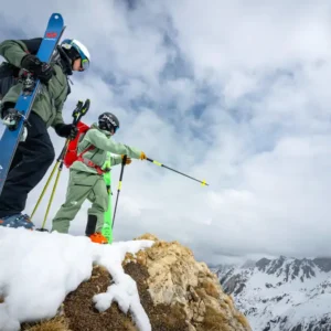 Two skiers with gear standing on snowy mountain ridge overlooking alpine peaks