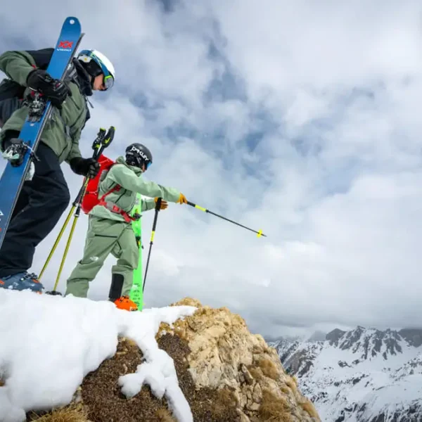 Two skiers with gear standing on snowy mountain ridge overlooking alpine peaks