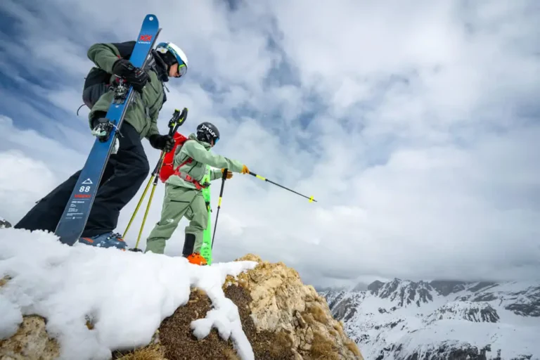 Two skiers with gear standing on snowy mountain ridge overlooking alpine peaks