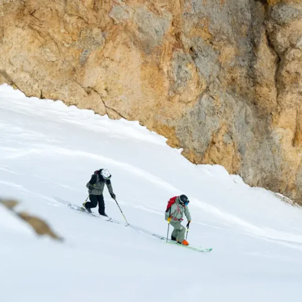Two skiers descending narrow snow slope beside rocky mountain wall