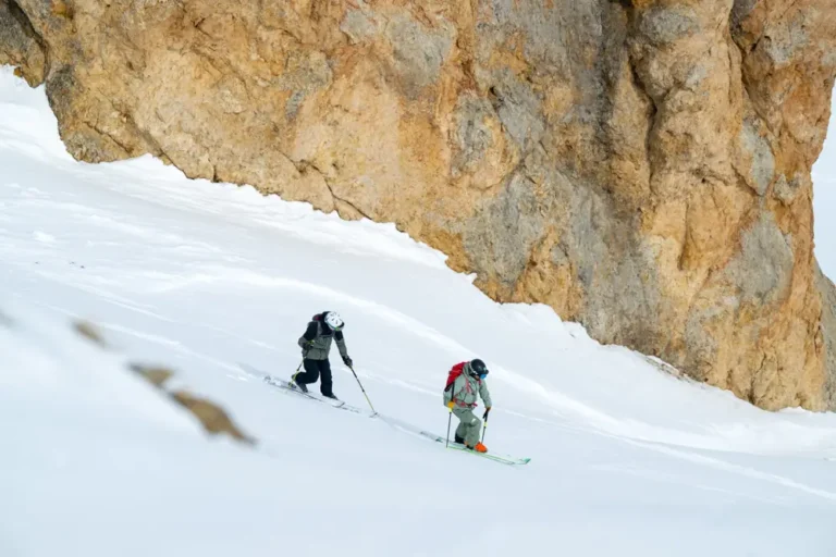 Two skiers descending narrow snow slope beside rocky mountain wall