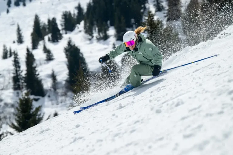 Female skier in green jacket carving turn with snow spray on forested slope